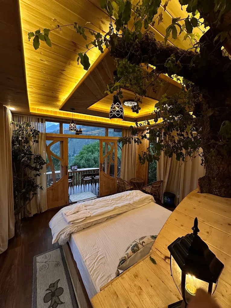 Deluxe Room from the foot of the bed: a tree rising beside it, carved timber doors and the valley beyond the balcony